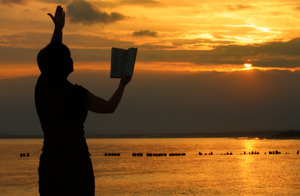 Woman stands praying from Bible at sunset